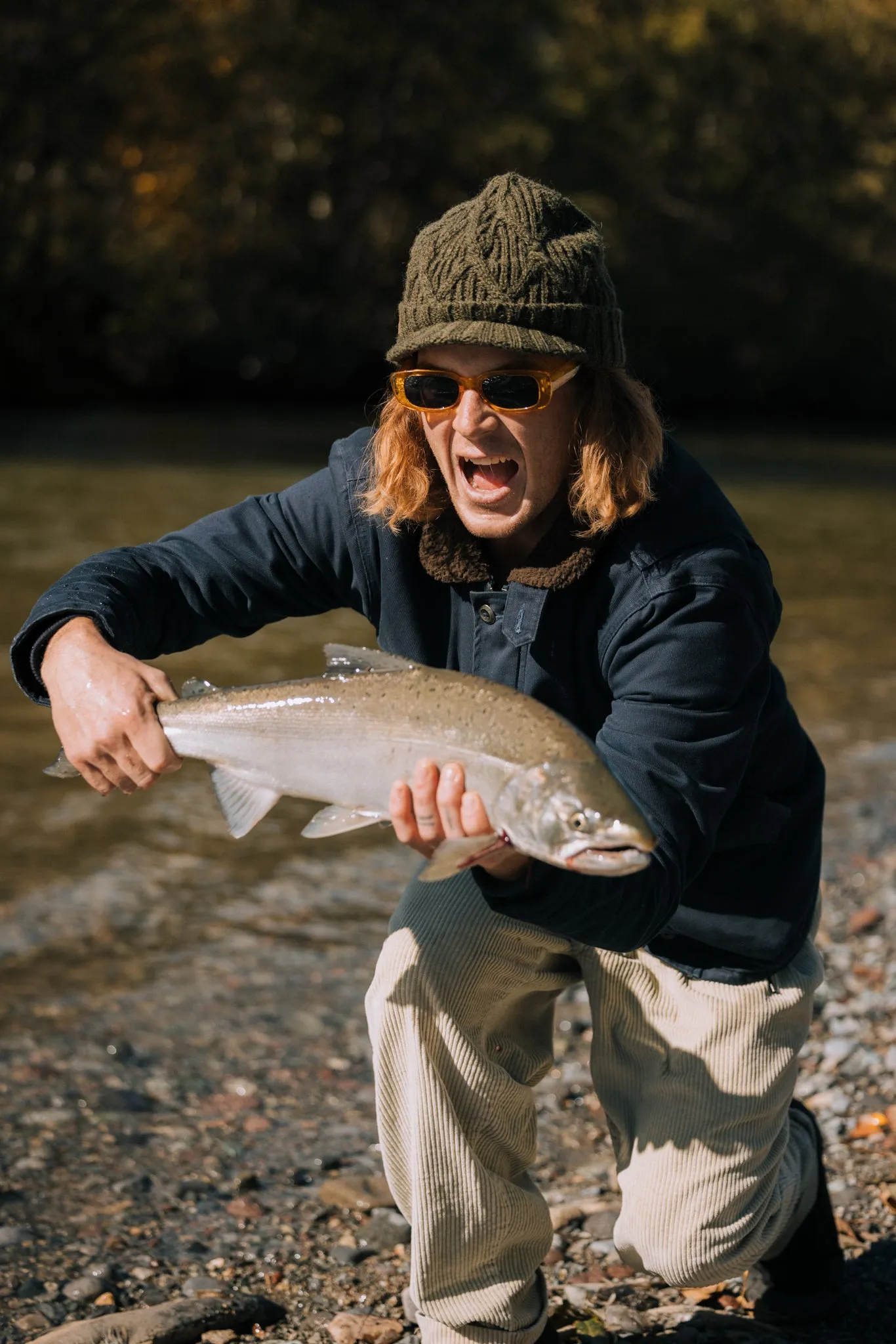 Angler in a Coal beanie laughing and holding a trout by a river