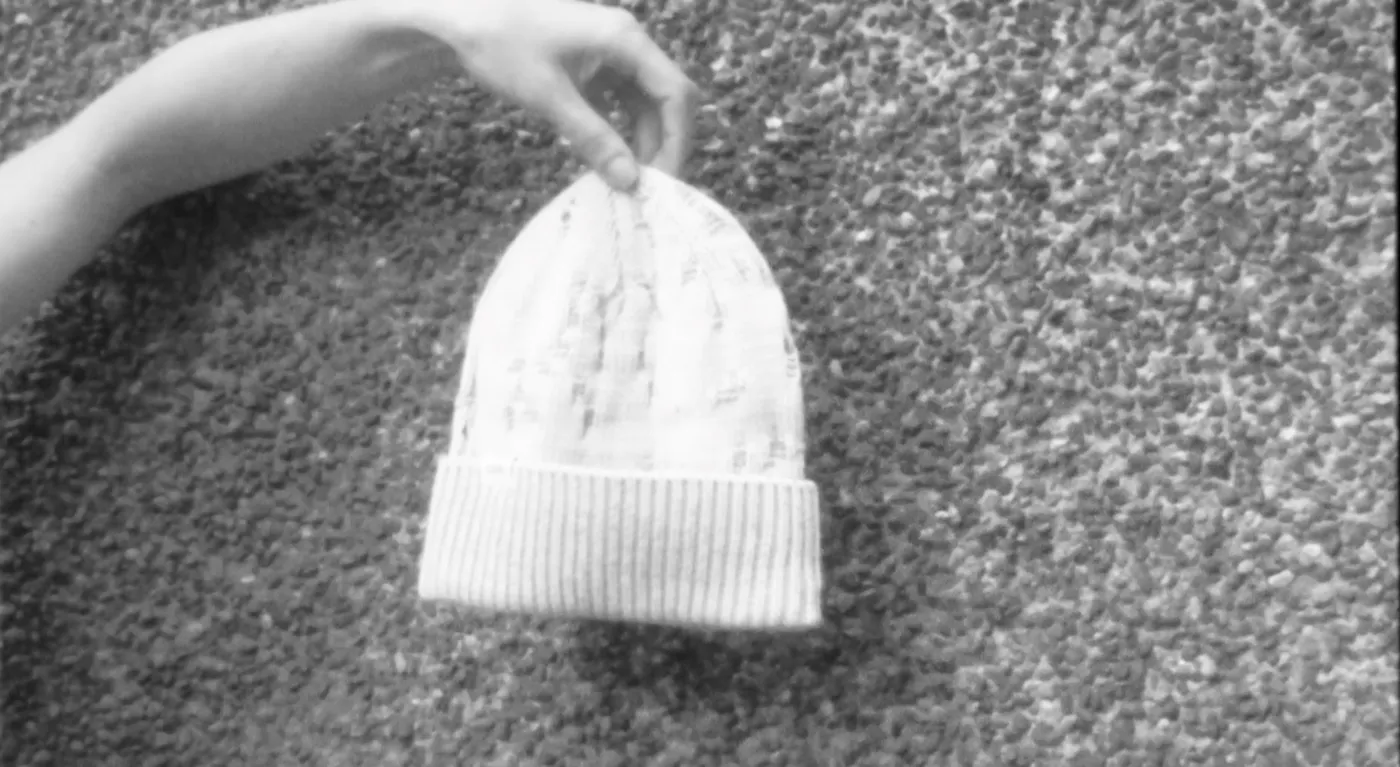 A hand holding a white knit beanie by its tip, the background in black and white
